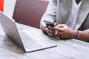 Pessoa digitando num celular, com um notebook em sua frente